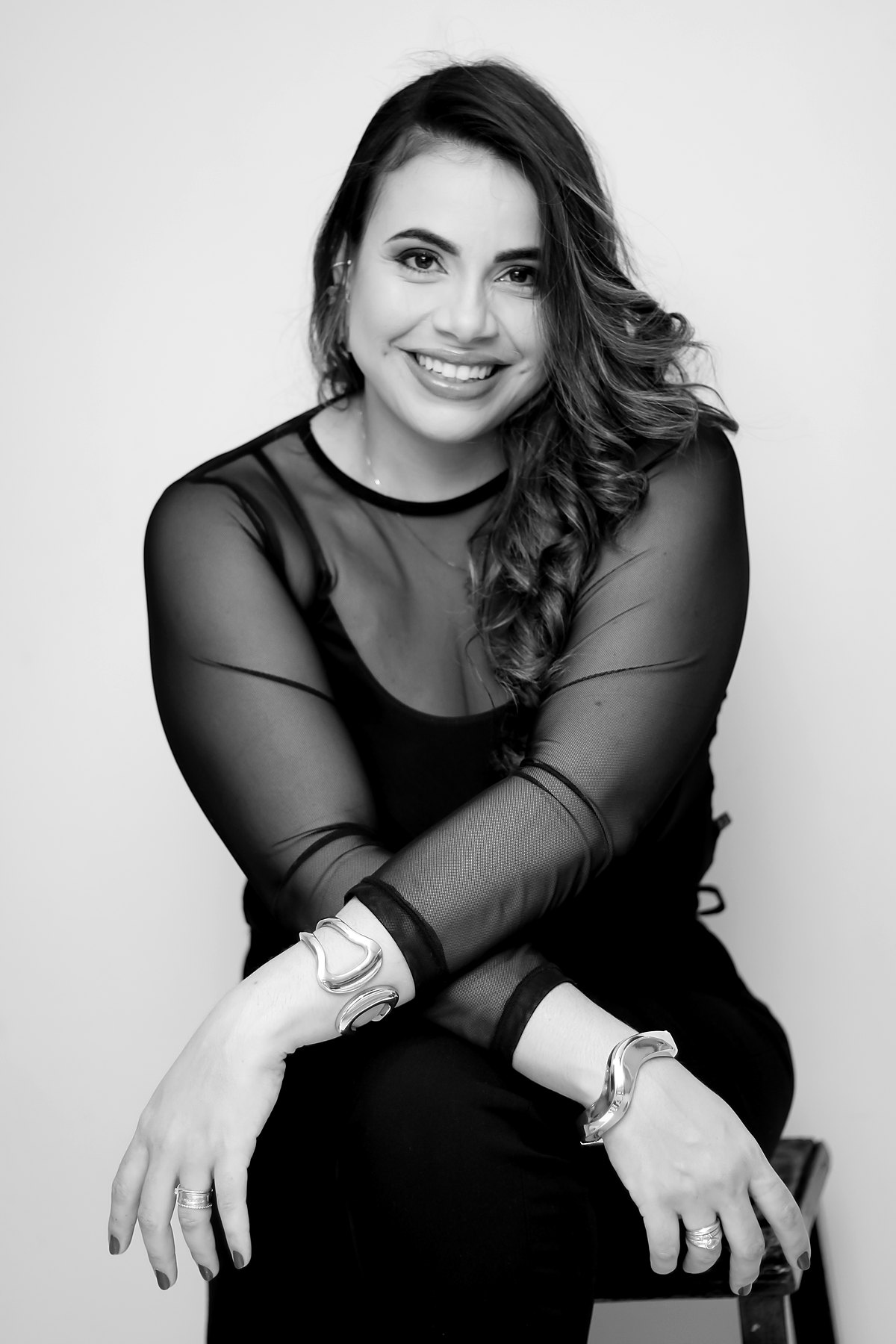 Retrato em preto e branco de Juliana Lopes Dias, sorrindo, sentada com os braços cruzados sobre os joelhos